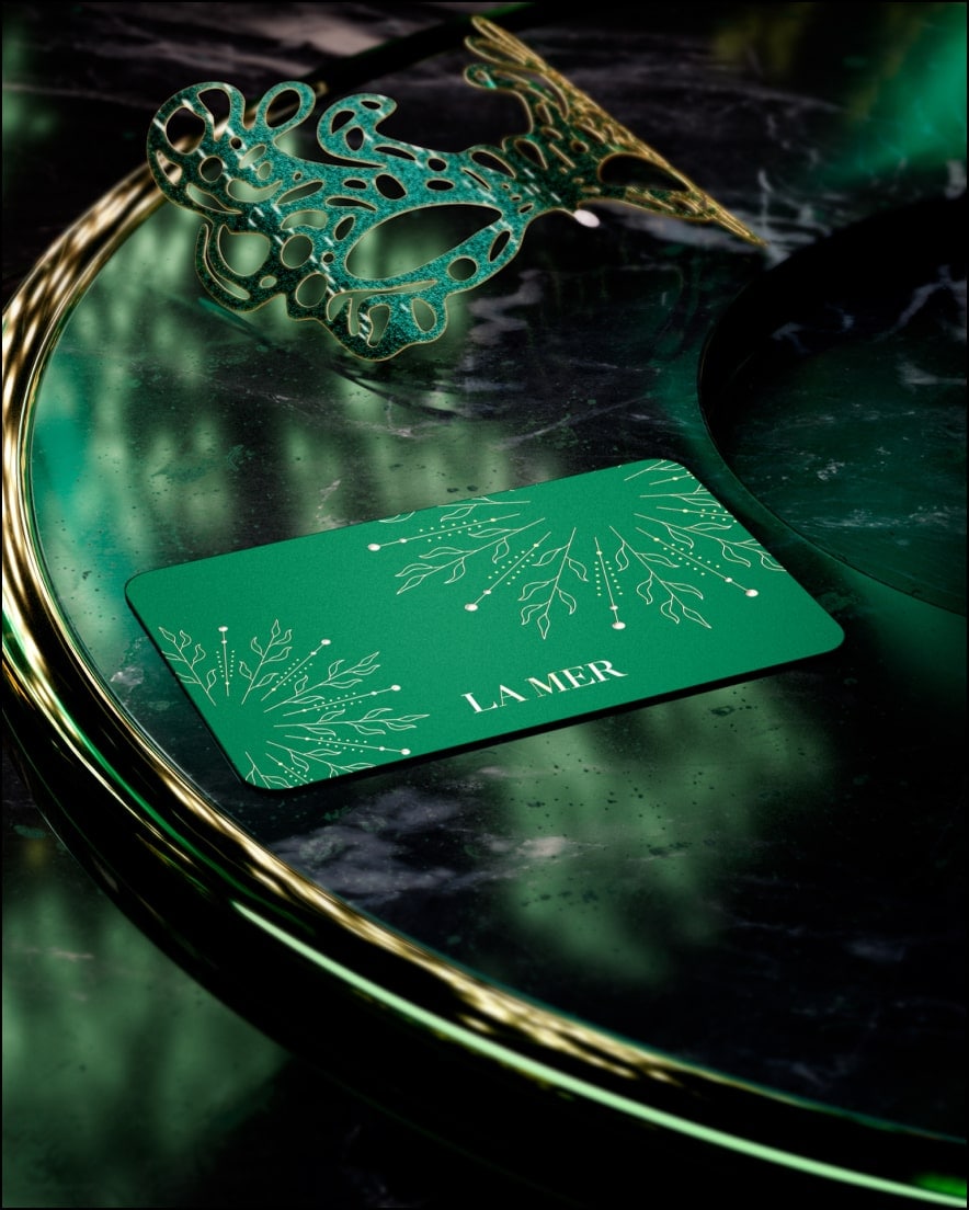 La Mer festive gift card on a glass table, nested next to a elegant, green masquerade mask